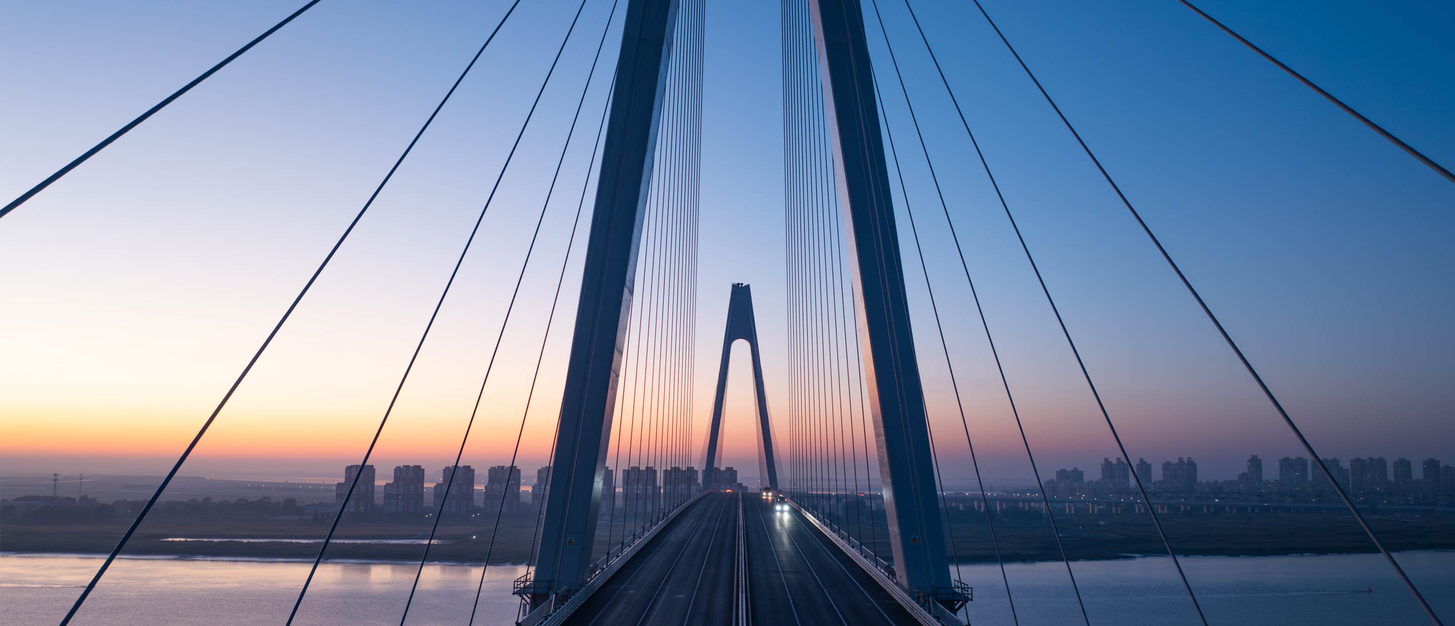 Modern cable-stayed bridge extending toward a city skyline at sunrise.